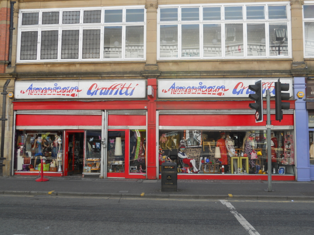 Eine Straßenszene mit einem Gebäude, Brettern, Schaufensterpuppen, Gläsern, einem Müllcontainer, einem Pfahl und Verkehrszeichen.