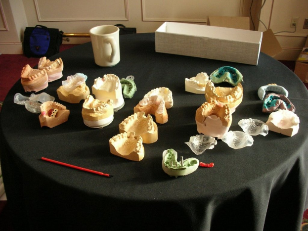 Ein schwarzer Tisch mit einem Becher, einem weißen Kasten und verschiedenen Gegenständen, positioniert vor einer Wand mit Taschen, Kartons und elektrischen Steckdosen, auf einem Teppichboden.