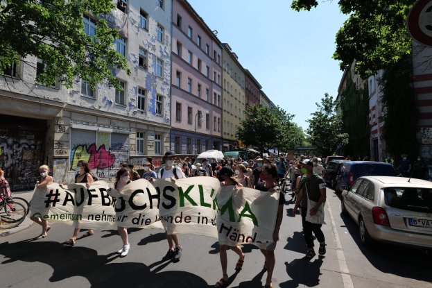 Eine Gruppe von Menschen marschiert auf einer Stadtstraße mit einem Banner, daneben geparkte Autos, ein Fahrradfahrer auf der linken Seite, Gebäude und Bäume im Hintergrund und ein Berliner Straßenschild.