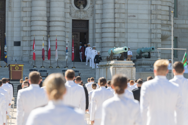 Gruppe von Menschen in weißen Marineuniformen, die vor einem Säulengebäude mit einer Tür stehen, mit Fahnen, einem Podium und Kanonen im Hintergrund.