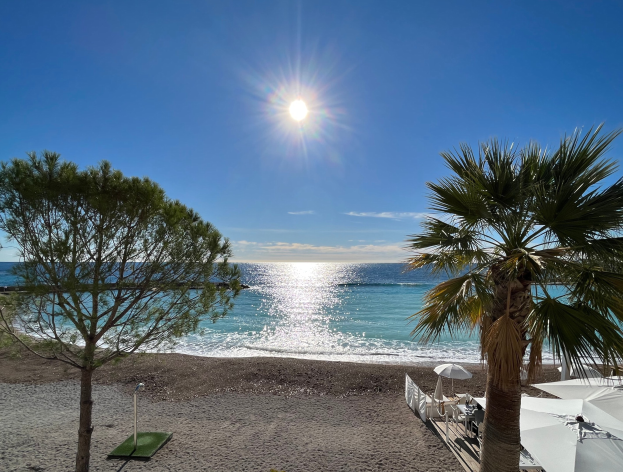Palmen und Sonnenschirme säumen einen sandigen Strand mit üppiger Vegetation unter einem strahlend blauen Himmel mit der Sonne im Hintergrund.