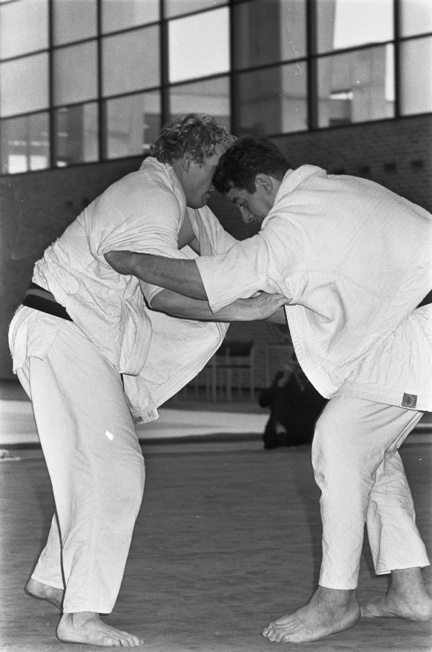 Zwei Männer in weißen Judo-Anzügen kämpfen intensiv auf einem Boden mit einer Wand und Glasfenstern im Hintergrund, dargestellt in Schwarz-Weiß.