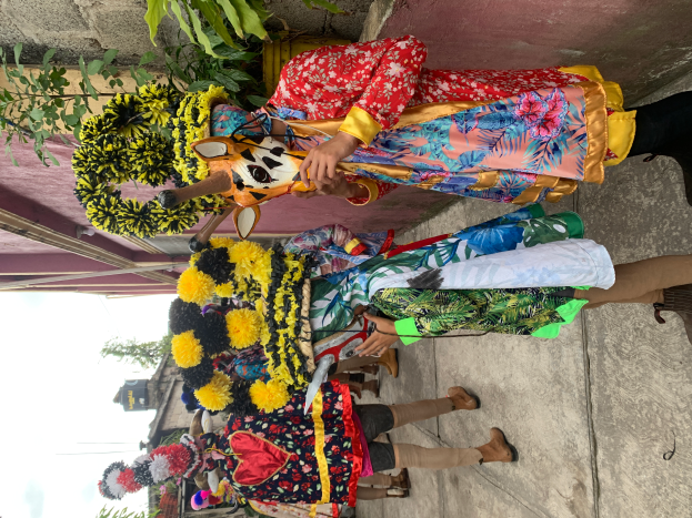 Eine Gruppe von Menschen in bunten Kostümen steht zusammen vor einem Gebäude, mit verschiedenen Gegenständen, Bäumen, Gebäuden und einem klaren blauen Himmel im Hintergrund bei einem Karnevalsumzug.