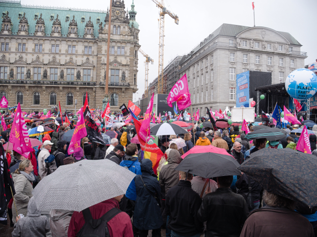 Eine große Gruppe von Menschen mit Schirmen und Plakaten nimmt an einer Frauen Demo in Berlin teil, mit Ballons und Stangen im Vordergrund und dem Himmel im Hintergrund.