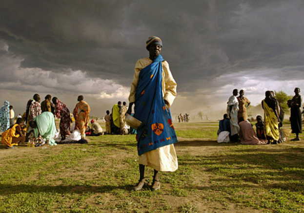 Eine Frau in einem blauen Kleid steht mit einem silbernen Gefäß in der Hand, umgeben von einer Gruppe sitzender und stehender Frauen unter einem bewölkten Himmel mit Bäumen im Hintergrund.
