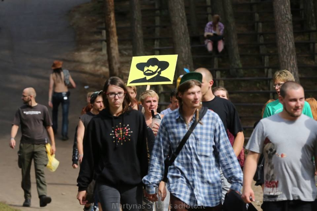 Eine Gruppe von Menschen geht einen Waldweg entlang, einige tragen Taschen und einer hält ein Schild mit einem Personenbild, mit Stufen und Bäumen im Hintergrund.