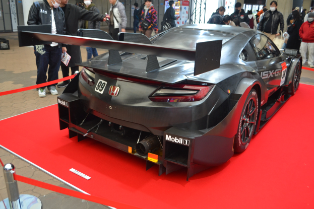 Ein roter und weißer Honda NSX GT3 Rennauto auf einer Indoor-Veranstaltung ausgestellt, umgeben von Menschen in bunten Kleidern und einigen mit Masken, mit einem roten Teppich daneben und Ausstellungsplakaten und Beleuchtung im Hintergrund.