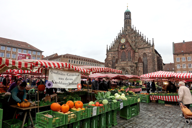 Ein belebter Markt in Nürnberg, Deutschland, mit farbenfrohen Obst- und Gemüsesorten auf dem Stand, Menschen mit Taschen und Zelten drumherum, mit Gebäuden und einem Kirchturm im Hintergrund unter einem sichtbaren Himmel.