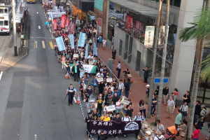 Eine große Gruppe von Menschen marschiert durch eine Straße in Hong Kong und hält Protestschilder und Banner in den Händen, wobei Bäume, Glasgebäude und Fahrzeuge auf beiden Seiten zu sehen sind.