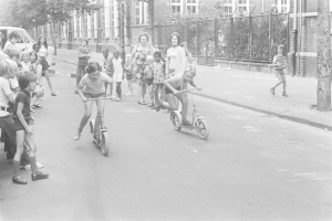 Schwarzes Bild von Kindern, die auf Rollern die Straße hinunterfahren, mit einem geparkten Auto auf der linken Seite, Gebäuden, Bäumen, Pfählen und einem Zaun im Hintergrund.