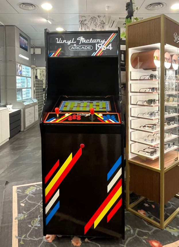 Ein Geschäft mit einem zentralen Retro-Arcade-Automaten, einem Glasvitrinenschrank auf der rechten Seite und Hintergrund-Elementen wie Schränken, einem Fernseher und anderen Gegenständen, mit Deckenleuchten, die eine retro-mäßige Atmosphäre schaffen.