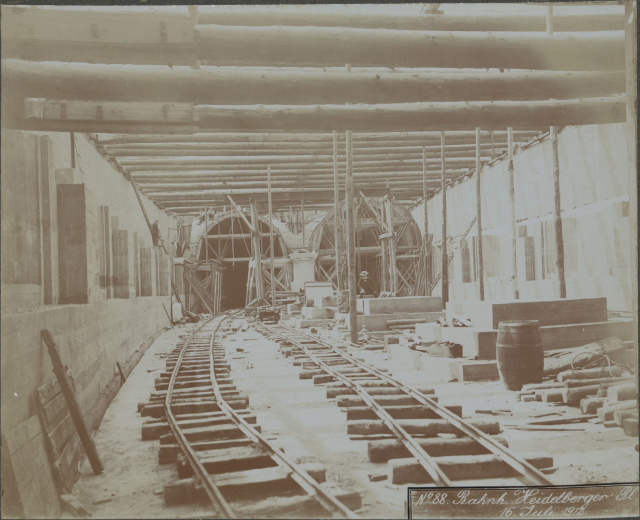 Schwarz-weiß-Foto einer Eisenbahnschiene in einem Gebäude unter Bau, mit verstreuten Holzgegenständen, Fässern, Wänden, Pfählen und Text am unteren Rand.