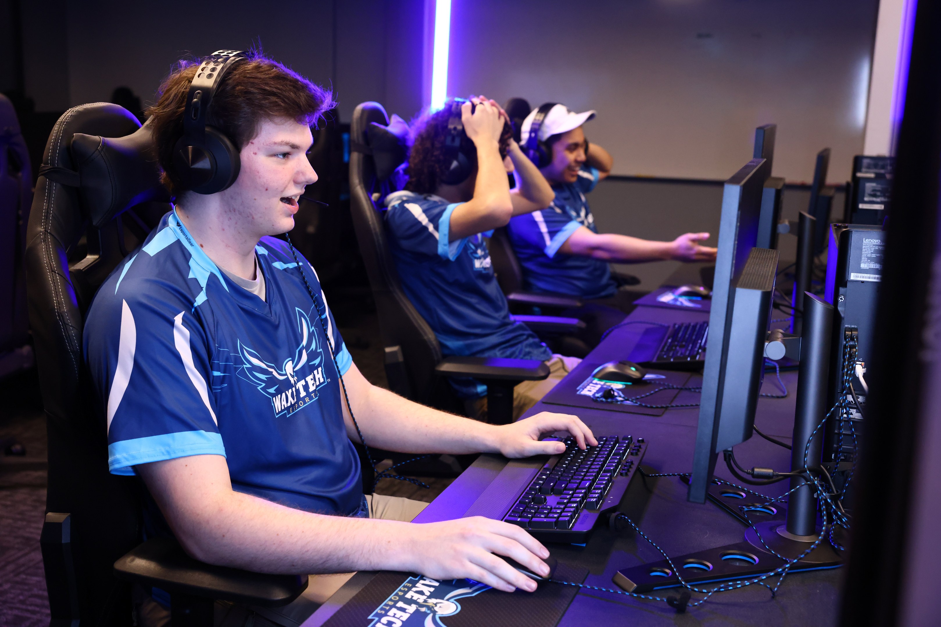 Gruppe junger M├Ąnner mit Headsets, die ein Videospiel auf Computern mit Monitoren, Tastaturen und M├Ąusen auf einem Schreibtisch spielen, mit beleuchteter Wand im Hintergrund.