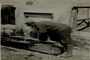 Ein altes Schwarz-Weiß-Foto eines Mannes in einer Mütze und Schuhen, der aus einem Brunnen trinkt, mit einer Wand, einem Fenster und einer Tafel mit Schrift hinter ihm.