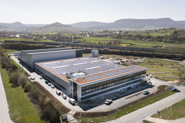 Luftaufnahme eines großen Industriegebäudes mit Solarpaneelen auf dem Dach, umgeben von Fahrzeugen, Bäumen, Gras und Strommasten, vor Bergen und einem klaren blauen Himmel.