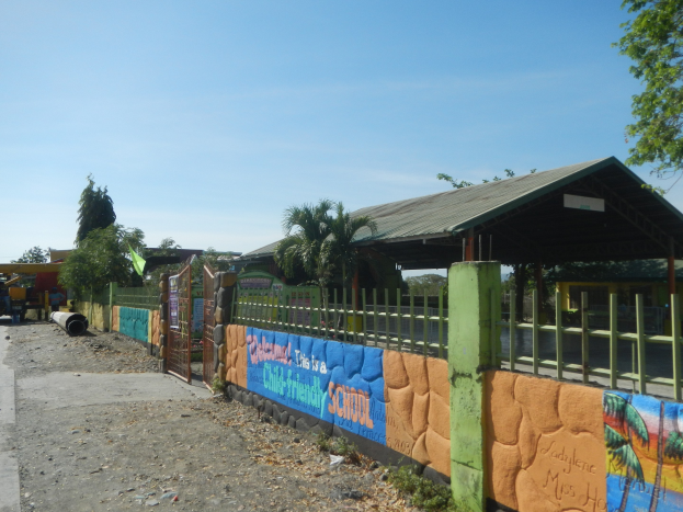 Schulgebäude mit Wandgemälde, umgeben von einem Zaun, neben einem Schuppen, Bäumen, Fahrzeugen, Strommasten, unter einem bewölkten Himmel.