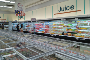 Ein hell erleuchteter Supermarktgang mit Flaschengetränken, verpackten Lebensmitteln und frischem Gemüse sowie einigen Kunden, die durch die Gänge schlendern und eine deckenmontierte Leuchttafel über ihnen.