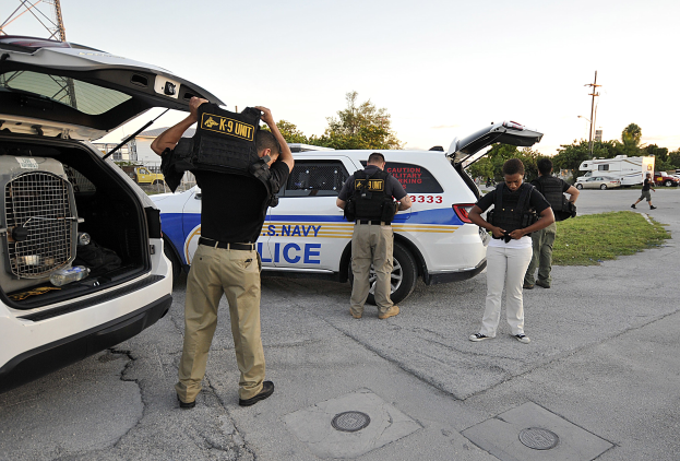 Polizeibeamte in Uniform mit K-9-Einheit-Taschen neben einem Polizeiwagen auf einer Straße mit Gras, Bäumen, Stromleitungen und einem Turm im Hintergrund bei klarem Himmel.
