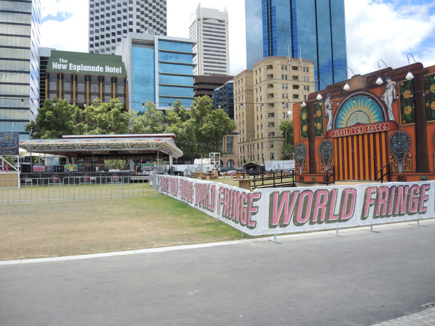 Ein Karnevals-Umzug in einer Stadtstraße mit hohen Gebäuden, Bäumen und einem bewölkten Himmel im Hintergrund, eine Straße mit einem Geländer und einem Banner mit Text am unteren Rand.