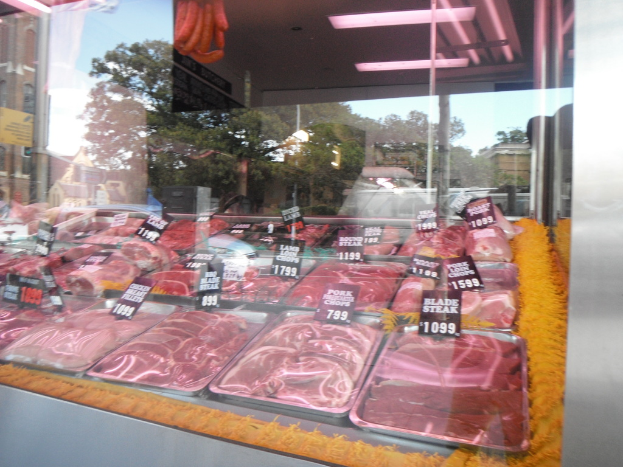 Eine Glasvitrine mit Tabletts voller Fleisch mit Preisschildern, mit Gebäuden und Bäumen durch die Glaswand und einer Decke oben.