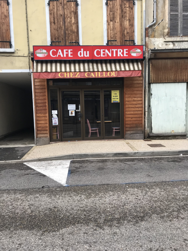 Ein zweistöckiges Gebäude mit einer 'Cafe du Centre'-Schilder an der Seite, das Fenster, eine Tür und eine Straße vorne mit einer Tafel zeigt, auf der Text steht.