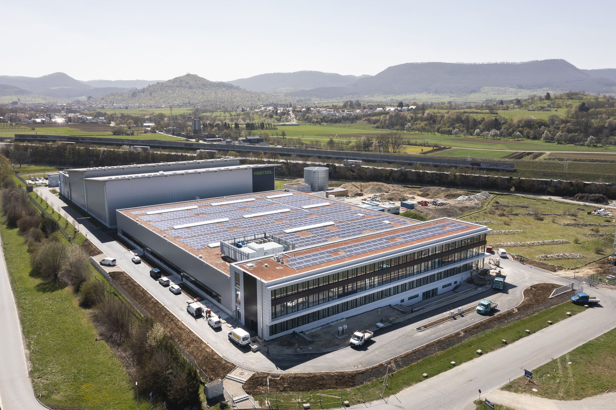 Luftaufnahme eines großen Gebäudes mit Solarmodulen auf dem Dach, umgeben von Fahrzeugen, Bäumen, Gras und Polen, mit Hügeln und einem klaren blauen Himmel im Hintergrund.