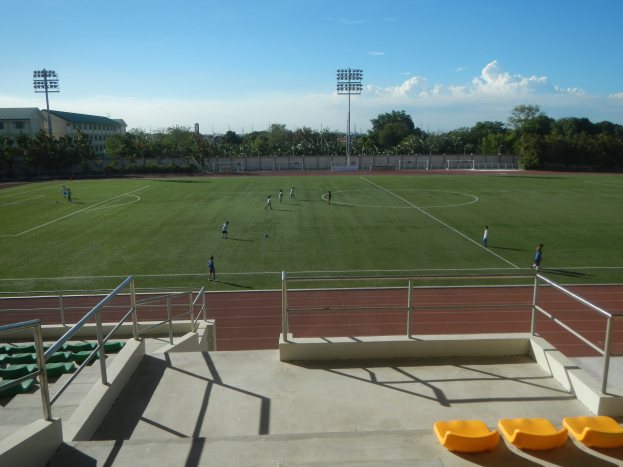 Ein Fuβballfeld mit spielenden Spielern, umgeben von Treppen, Geländern und gelben Objekten am Boden sowie Bäumen, Laternen, Gebäuden und einem bewölkten Himmel im Hintergrund.