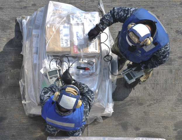 Zwei Männer in blauen Uniformen und Helmen arbeiten an einem großen Drohnenmodell, jeder hält Werkzeuge, mit einer Plastikhülle auf dem Boden.