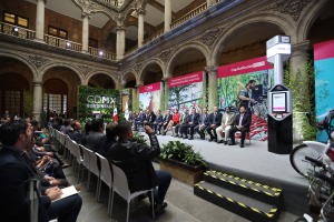 Eine Gruppe von Menschen, die in Stühlen sitzen und einem Mann auf einer Bühne mit einem Mikrofon zuhören, mit Fahrrädern, die rechts geparkt sind, und verschiedenen Elementen wie Plakaten, Pflanzen, Säulen, Geländern, Lampen, Fenstern und einer Wand im Hintergrund.
