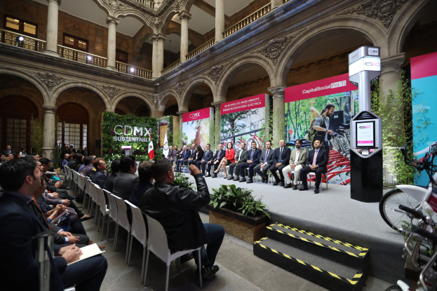Eine Gruppe von Menschen, die in Stühlen sitzen und einem Mann auf einer Bühne mit einem Mikrofon zuhören, mit Fahrrädern, die rechts geparkt sind, und verschiedenen Elementen wie Plakaten, Pflanzen, Säulen, Geländern, Lampen, Fenstern und einer Wand im Hintergrund.