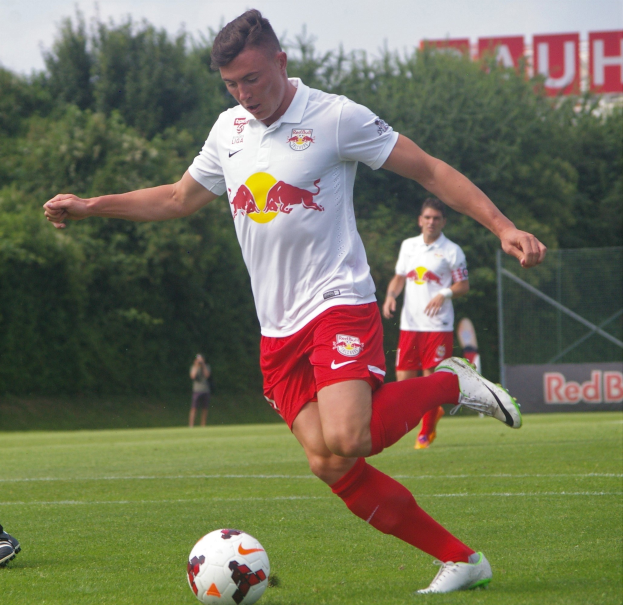 Ein professioneller Fußballspieler schießt den Ball auf einem Rasenfeld, wobei "RB Leipzig" im Hintergrund unter einem klaren blauen Himmel zu sehen ist.