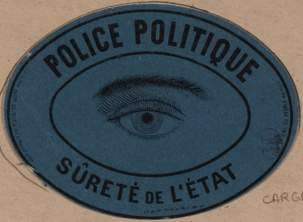Brauner Karton mit einem blauen Aufkleber mit der Aufschrift "Polizei Politique Sûreté de l'État" und einem zentralen Auge-Symbol.