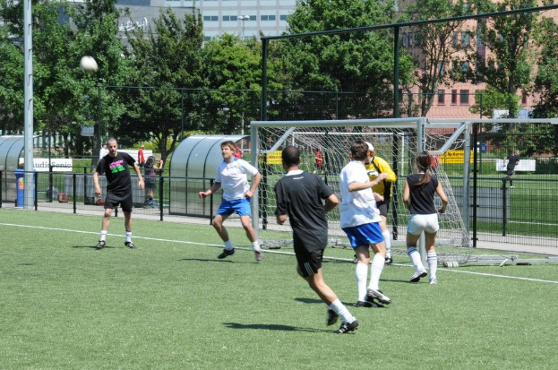 Menschen in Sportbekleidung beim Fußballspielen auf einem Feld mit einem Zaun und einem Tor, einem Pfosten und einem Ball oben links und Bäumen und Gebäuden im Hintergrund.