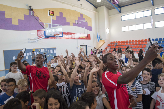 Kinder auf einem Basketballfeld mit Handys stehend, mit einer Pinnwand, Uhr, Torstange, Basketballkorb, Deckenbeleuchtung, Stühlen und Fenstern im Hintergrund, was auf ein Basketballcamp hinweist.