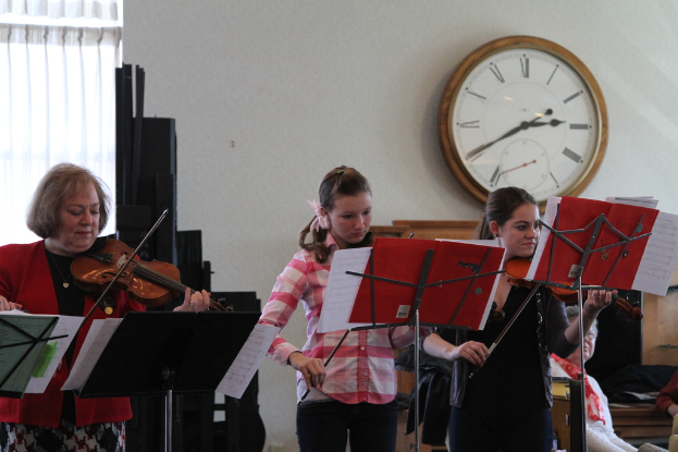 Drei Frauen auf der Bühne mit Geigen stehend und auf Musikständern spielend, während eine Gruppe von sitzenden Frauen im Vordergrund zuhört; eine Uhr hängt an einer weißen Wand im Hintergrund, mit einem weißen Vorhang in der linken Ecke.