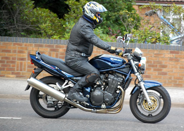Eine Person in einer schwarzen Jacke fährt mit dem Fahrrad auf einer Straße, trägt einen Helm und eine Schutzbrille, mit einem Gehweg, einer Wand, Bäumen und einem Haus im Hintergrund.