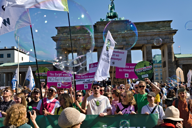 Eine Menschenmenge steht vor dem Brandenburger Tor in Berlin, Deutschland, einige tragen Mützen und Schutzbrillen, halten Fahnen und Schilder, darunter eines im Vordergrund, während Gebäude, ein Tor mit einer Statue und ein Himmel mit Seifenblasen im Hintergrund zu sehen sind.