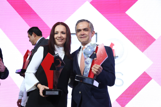 Zwei Personen, die auf einer Bühne strahlen und Trophäen halten, mit einem Bildschirm im Hintergrund.