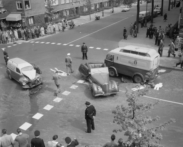 Schwarz-weiß-Szene eines Autounfalls an einer Straßenkreuzung mit einem Van, anderen Fahrzeugen und Menschen drumherum, mit Gebäuden, Bäumen, Pfählen und Texttafeln im Hintergrund.