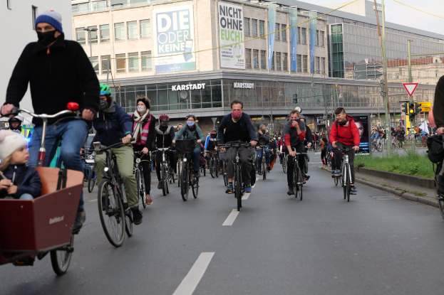 Eine Gruppe von Menschen, die auf Fahrrädern eine Straße entlangfahren, mit einem Kind in einem Wagen auf der linken Seite, Gebäuden und Bannern im Hintergrund unter einem klaren blauen Himmel.