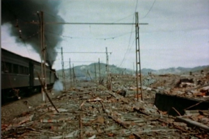 Ein Zug fährt durch ein beschädigtes Gebiet mit aufsteigendem Rauch, umgeben von Strommästen und -drähten, Hügel und einem klaren Himmel im Hintergrund und verstreuten Schrottstücken am Boden.