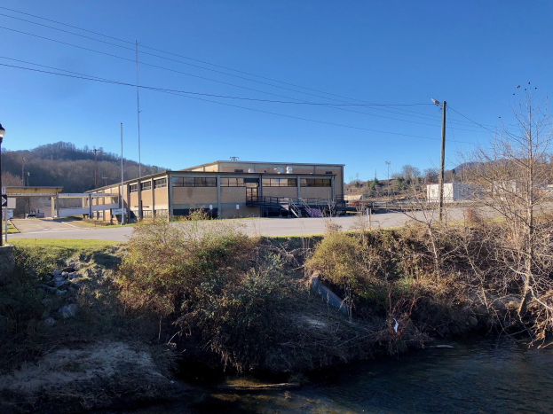 Ein Fluss fließt durch ein grünes Feld neben einem Gebäude in der Nähe von Bäumen, Pflanzen, Gras, Steinen, Straßenmasten, Straßenlaternen, Strommasten, Kabeln, Hügeln und einem klaren blauen Himmel, wobei das Gebäude als Standort für eine Wasseraufbereitungsanlage geplant ist.