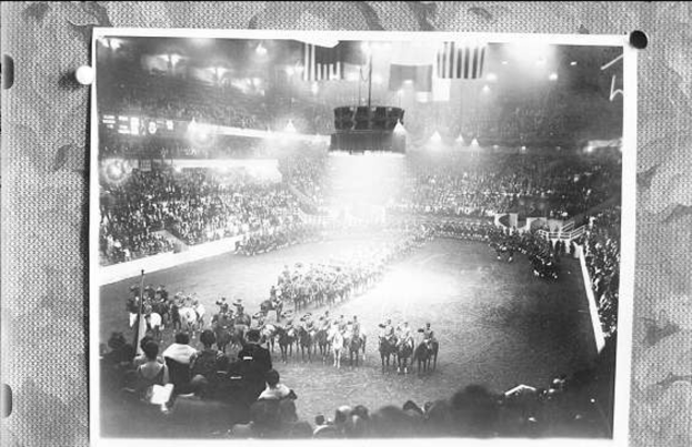 Ein Schwarz-Weiß-Foto einer großen Menschenmenge in einem Stadion, mit einigen Menschen auf Pferden und anderen, die stehen, eine Bühne mit Flaggen und Lichtern in der Mitte und Text unten.