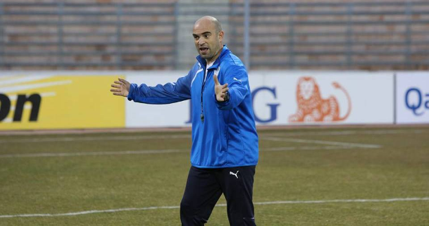 Ein Mann in einer blauen Jacke steht auf einem Fußballfeld, umgeben von Tafeln mit Text und Logos, entschlossen.