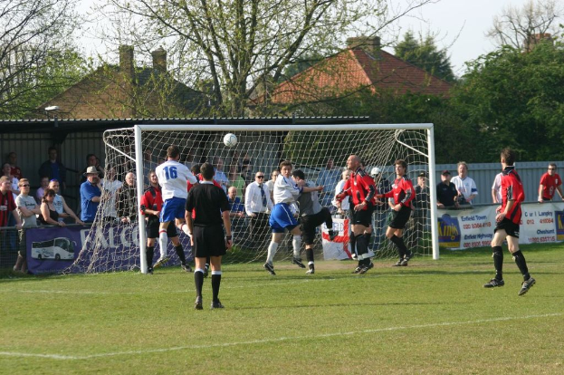 Spieler spielen Fußball auf einem Feld mit einem Tor und Zuschauern dahinter, mit Bäumen und Häusern im Hintergrund.