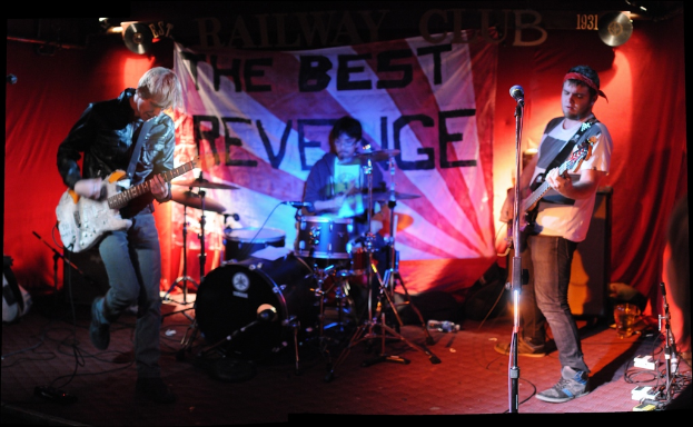 Zwei Personen vor einem Mikrofon mit Gitarren stehend, mit einem Mann mit Brille dahinter am Schlagzeug, vor einem Hintergrund mit flexiblen Schallplatten und CDs.
