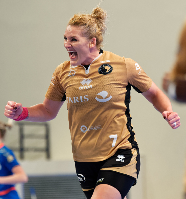 Eine Frau in braunem T-Shirt und schwarzen Shorts rennt auf einem Futsal-Platz, feiert mit einem triumphierenden Gesichtsausdruck, während zwei andere Frauen im Hintergrund unscharf sind.