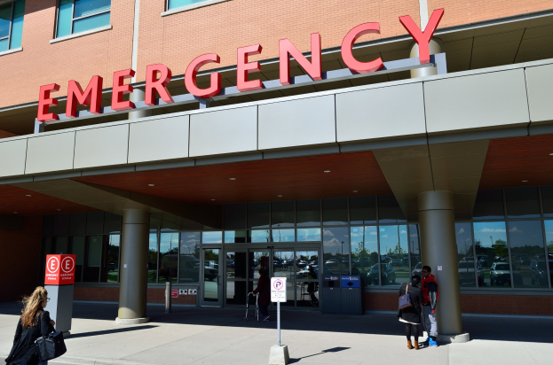 Gruppe von Menschen, die auf ein Notaufnahmelokal mit Glaswänden, Säulen und einem Namensschild zusteuern, begleitet von Schildern und einem Müllcontainer.