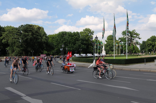 Eine Gruppe von Menschen, die auf Fahrrädern eine von Bäumen gesäumte Straße mit Laternenmasten und Flaggenmasten entlangfahren, während der Pariser Radtour 2016, mit Fahrzeugen und bewölktem Himmel im Hintergrund.