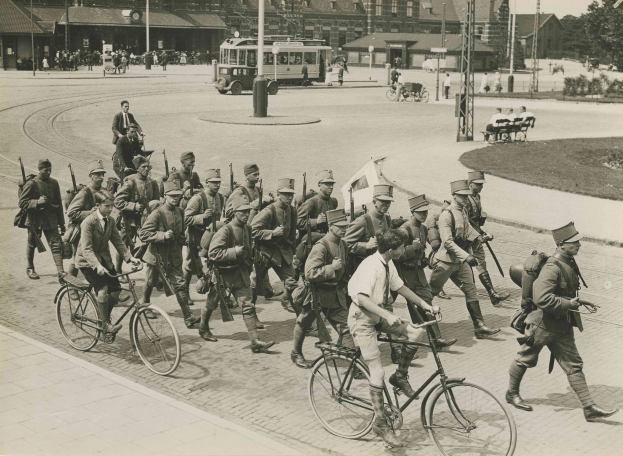 Schwarz-weißes Bild einer Gruppe von Männern, die auf Fahrrädern eine Straße entlangfahren, einige halten Waffen in den Händen, mit Gebäuden, Fahrzeugen und Bäumen im Hintergrund.